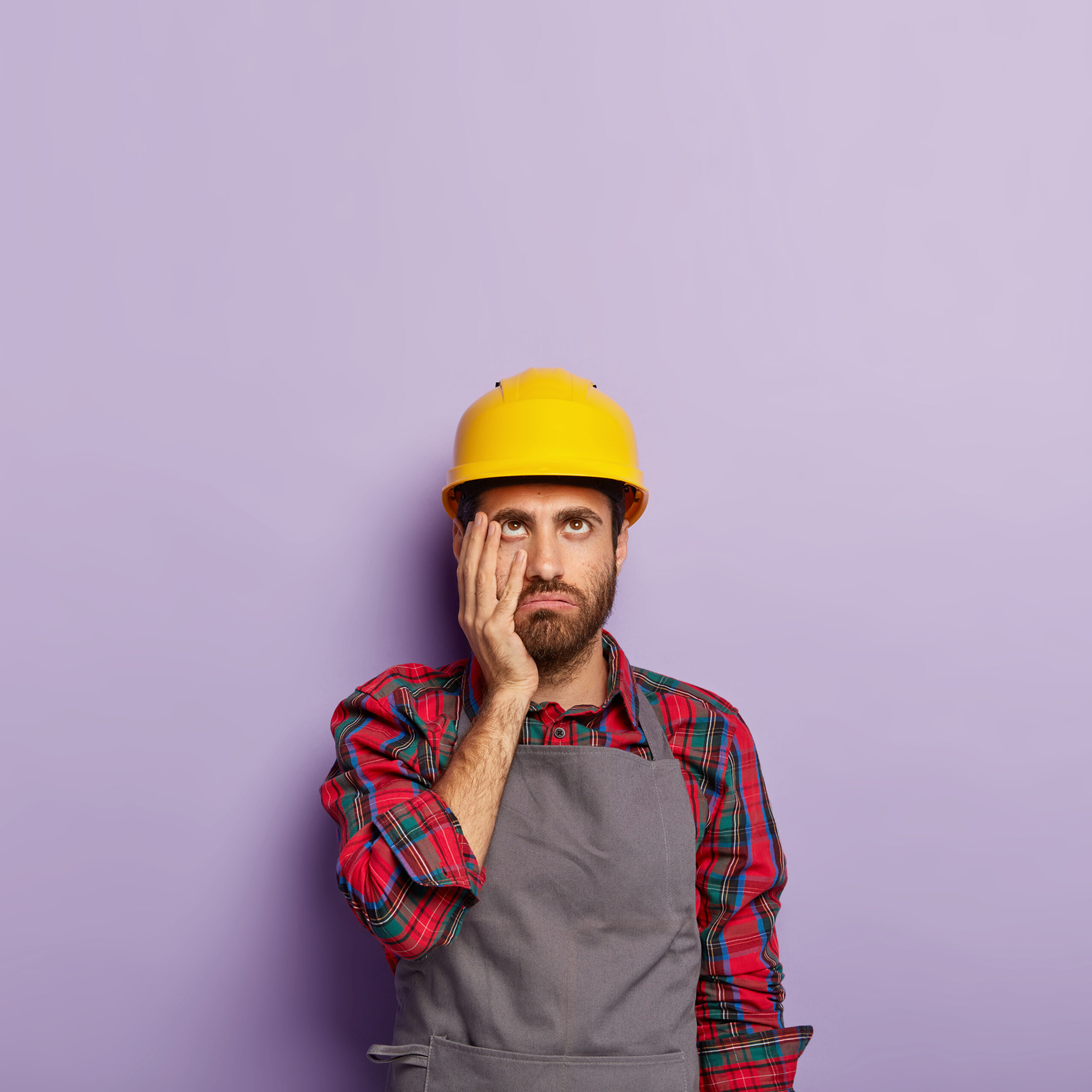 Tired technician touches cheek, focused up, has to repair one more item, wears hardhat on head, casual apron, busy with duties, isolated on purple wall. Experienced construction worker indoor